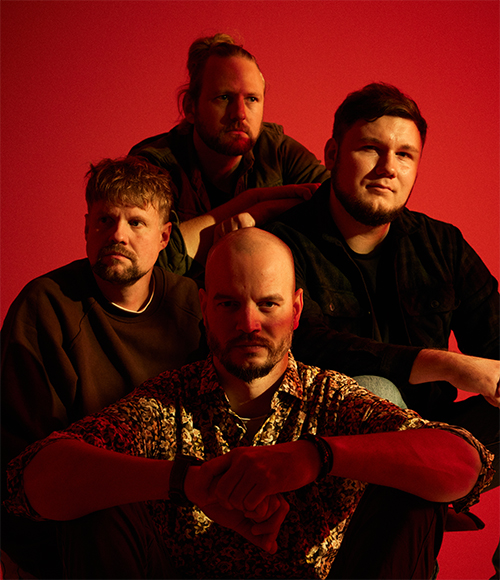 Bandfoto. Alle vier Band Mitglieder sitzennah beieinander. Sie sind nur bis zur Hüfte zu sehen. Lowlight mit rot-orangenem Hintergrund.